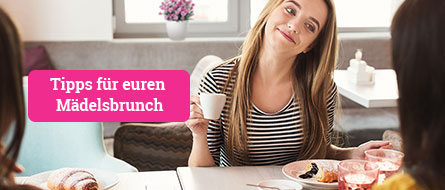 Eine junge Frau sitzt offensichtlich mit ihren Freundinnen am Frühstückstisch, sie lächelt die junge Frau ihr gegenüber an, von der nur ein Teil des Hinterkopfes zu sehen ist. In der Hand hält sie eine Kaffeetasse, vor ihr steht ein Teller mit einem Schoko-Croissant, im Hintergrund ist unscharf eine Pink Kisses Mininelke im Topf audf der Fensterbank zu erkennen.